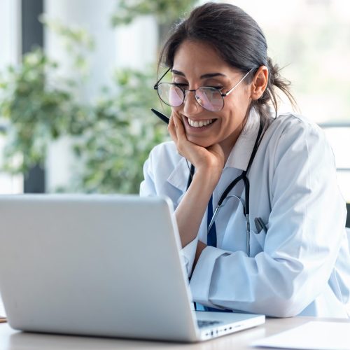Eine lächelnde Ärztin mit dunklem Haar und Brille sitzt an einem Schreibtisch und macht am Laptop eine digitale Weiterbildung in der Medizininformatik.