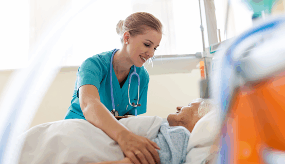 © istockphoto.com/PIKSEL 1157106508 Eine Krankenschwester in türkisfarbener Arbeitskleidung beugt sich lächelnd über eine ältere Patientin, die in einem Krankenhausbett liegt. Die Krankenschwester legt beruhigend ihre Hand auf den Arm der Patientin.