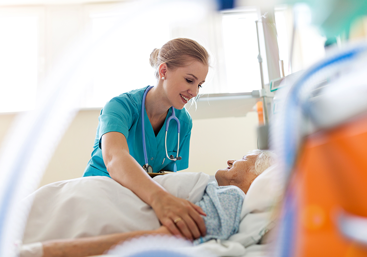 Eine Krankenschwester aus der regionalen Patientenversorgung beugt sich lächelnd über eine ältere Patientin, die in einem Krankenhausbett auf der Intensivstation liegt.