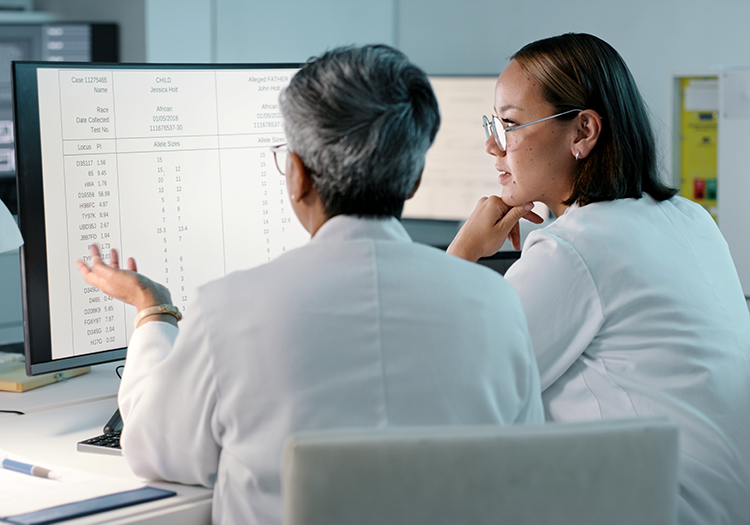Zwei Wissenschaftlerinnen in weißen Kitteln sitzen vor einem Computerbildschirm und analysieren Patientendaten aus der regionalen Routineversorgung.