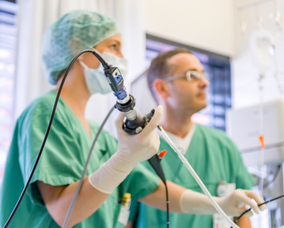 Zwei Ärzte in grüner OP-Kleidung und Masken führen eine medizinische Untersuchung mit einem Endoskop durch. Der Fokus liegt auf der Hand der Ärztin, die das Endoskop hält. Im Hintergrund ist ein weiterer Arzt unscharf zu erkennen. Diese Szene zeigt die Arbeit in der Intensivmedizin, wo moderne Technik und präzises Handeln essenziell sind.