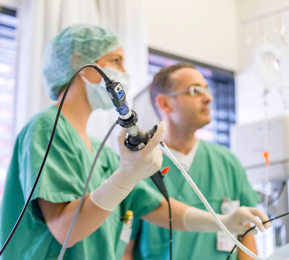 Zwei Ärzte in grüner OP-Kleidung und Masken führen eine medizinische Untersuchung mit einem Endoskop durch. Der Fokus liegt auf der Hand der Ärztin, die das Endoskop hält. Im Hintergrund ist ein weiterer Arzt unscharf zu erkennen. Diese Szene zeigt die Arbeit in der Intensivmedizin, wo moderne Technik und präzises Handeln essenziell sind.