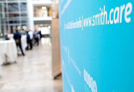 Eine Nahaufnahme einer Plakatwand auf dem SMITH-Kongress 2022. Im Hintergrund unterhalten sich Menschen an Stehtischen.