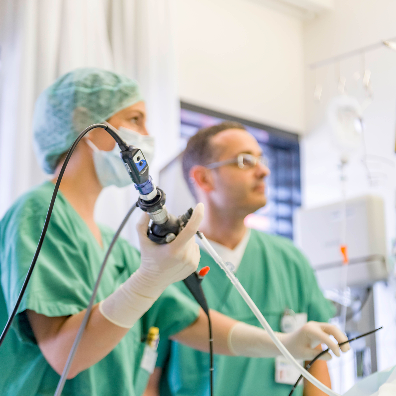 Zwei Ärzte in grüner OP-Kleidung und Masken führen eine medizinische Untersuchung mit einem Endoskop durch. Der Fokus liegt auf der Hand der Ärztin, die das Endoskop hält. Im Hintergrund ist ein weiterer Arzt unscharf zu erkennen. Diese Szene zeigt die Arbeit in der Intensivmedizin, wo moderne Technik und präzises Handeln essenziell sind.